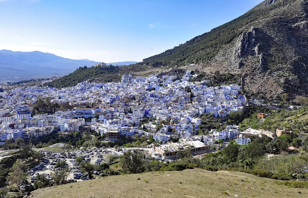 Vista do alto de Chefchaouen, com casas brancas e azuis encostadas numa montanha coberta de pinheiros e um estacionamento ao pé da cidade.