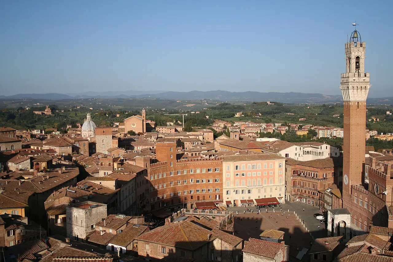 Vue d’en haut de Sienne, avec la Piazza del Campo en forme de coquille, la Torre del Mangia à droite, des toits de tuiles et des collines au fond.
