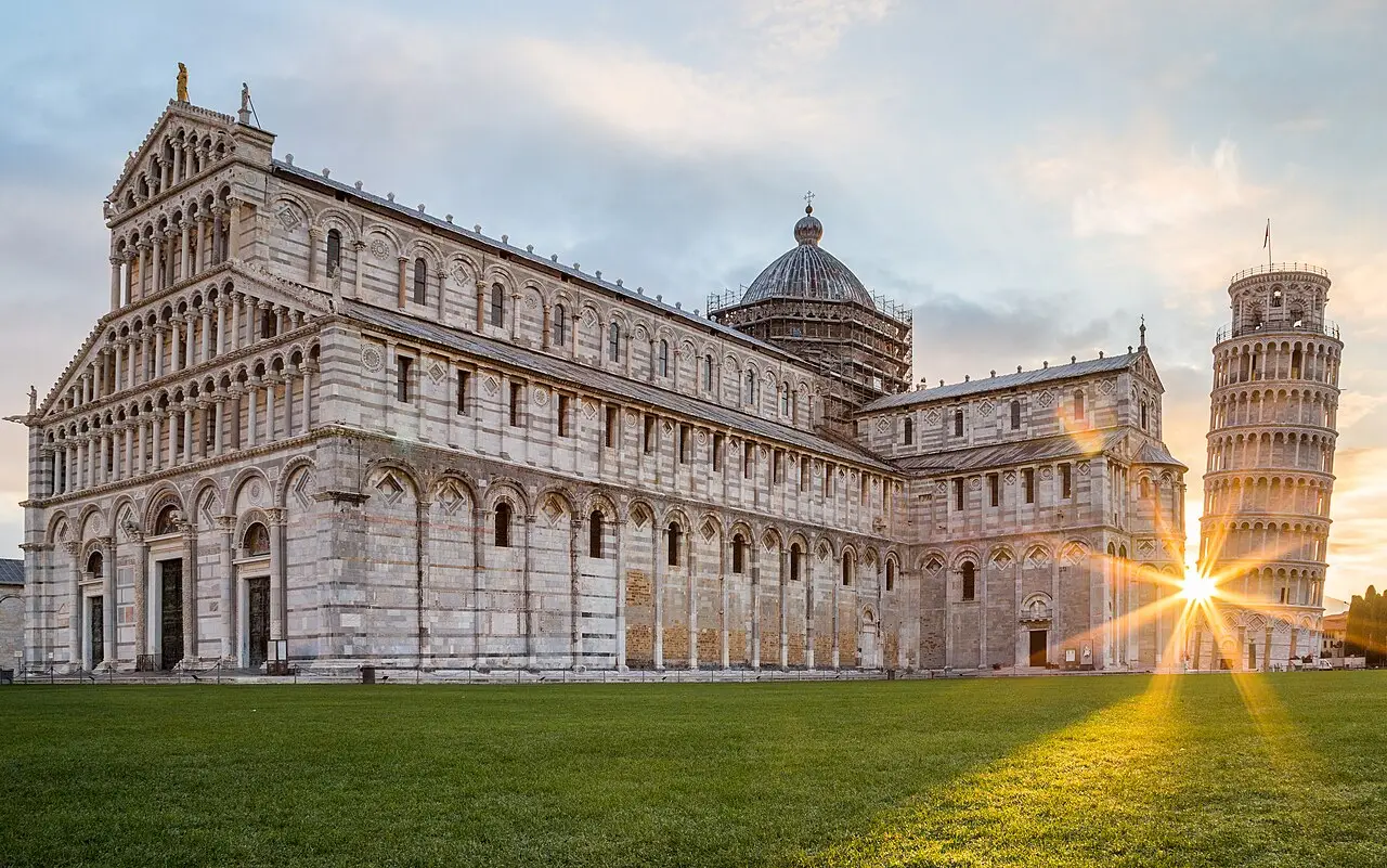 Duomo de Pisa de mármore listrado e a Torre inclinada à direita, com um raio de sol ao nascer do dia sobre o gramado da praça.