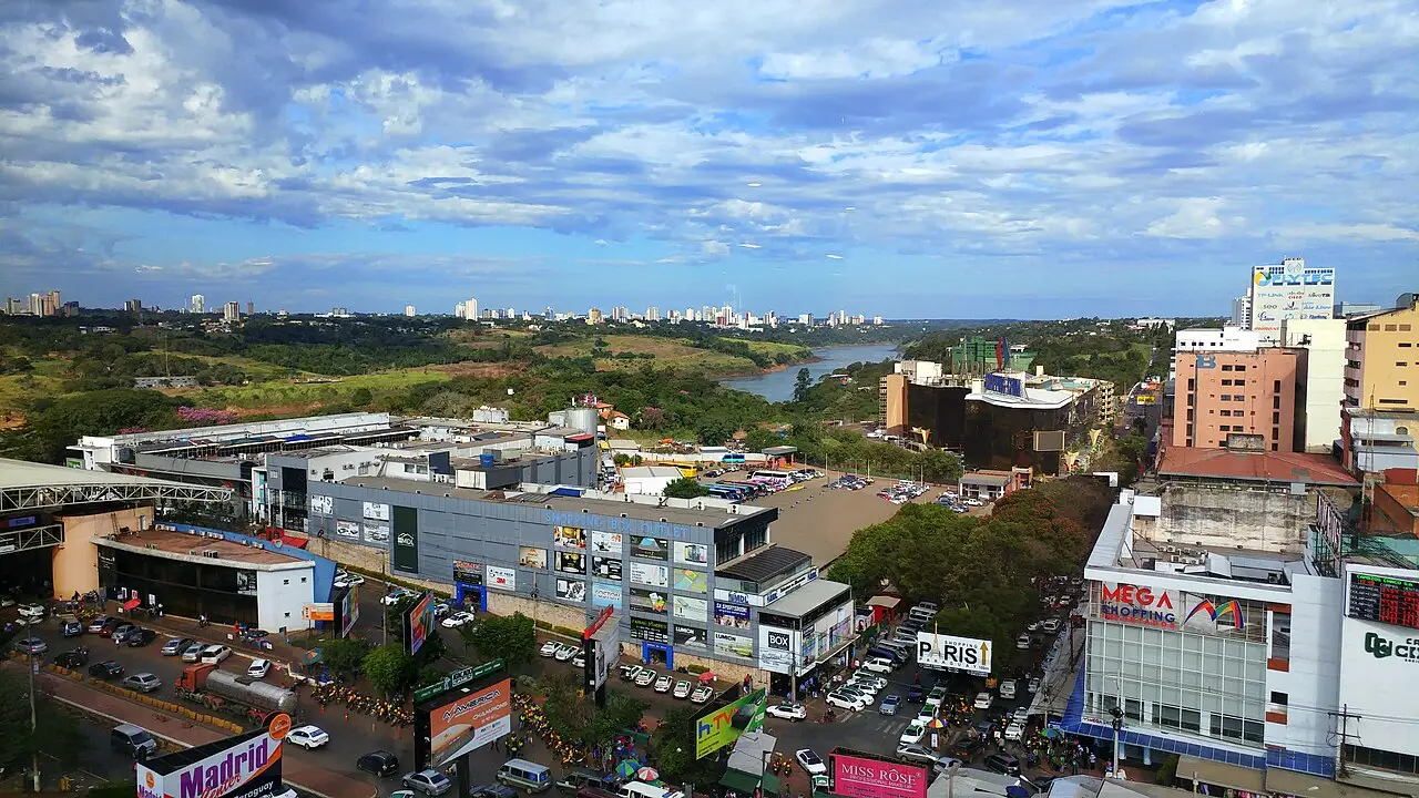 Lojas e prédios à beira de uma via, com o rio e mais cidade ao fundo.