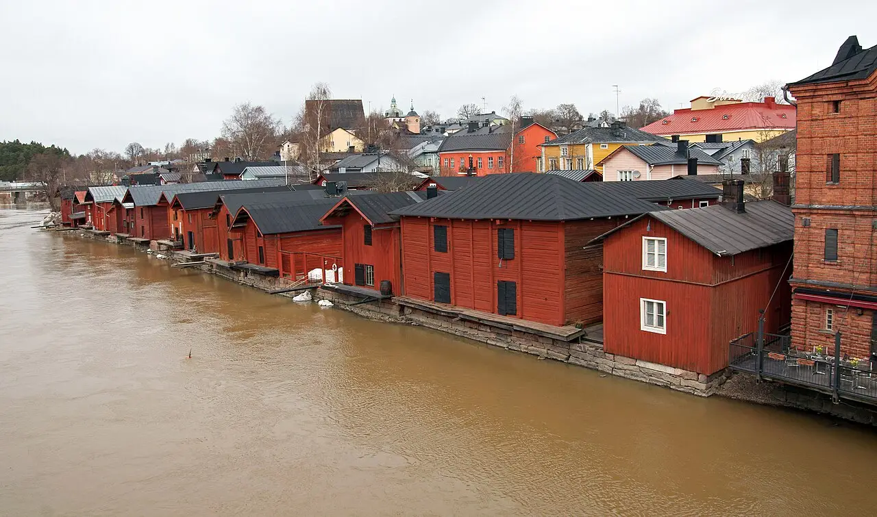 Armazéns de madeira vermelhos enfileirados à beira do rio de água marrom em Porvoo, com casas coloridas e um prédio de tijolos ao fundo.