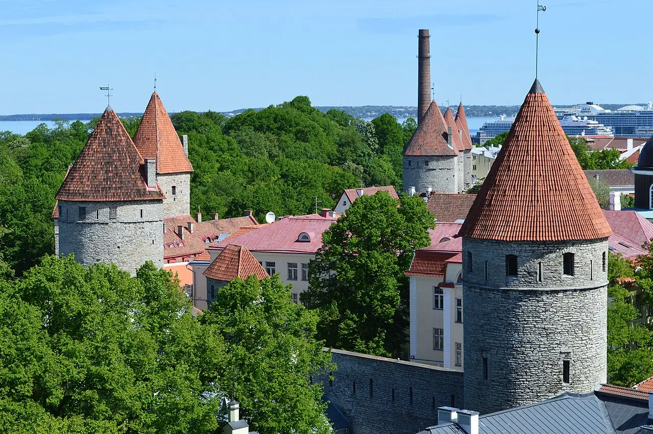 Torres redondas de pedra com telhados cônicos de telha vermelha na muralha de Tallinn, entre árvores verdes, com um navio de cruzeiro ao fundo.