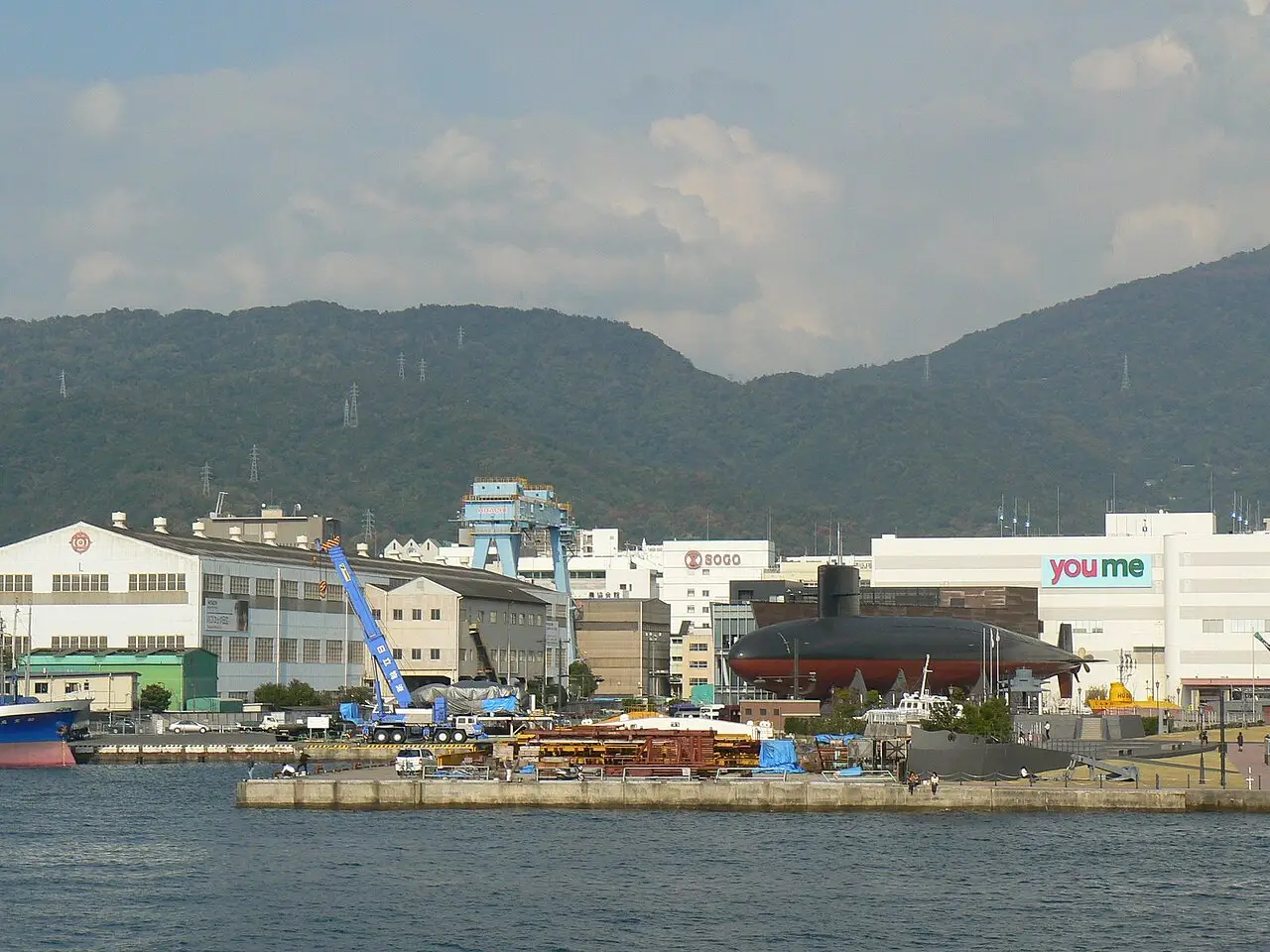 Porto de Kure com um submarino exposto em terra, galpões e um guindaste de estaleiro, um shopping e montanhas cobertas de mata ao fundo.
