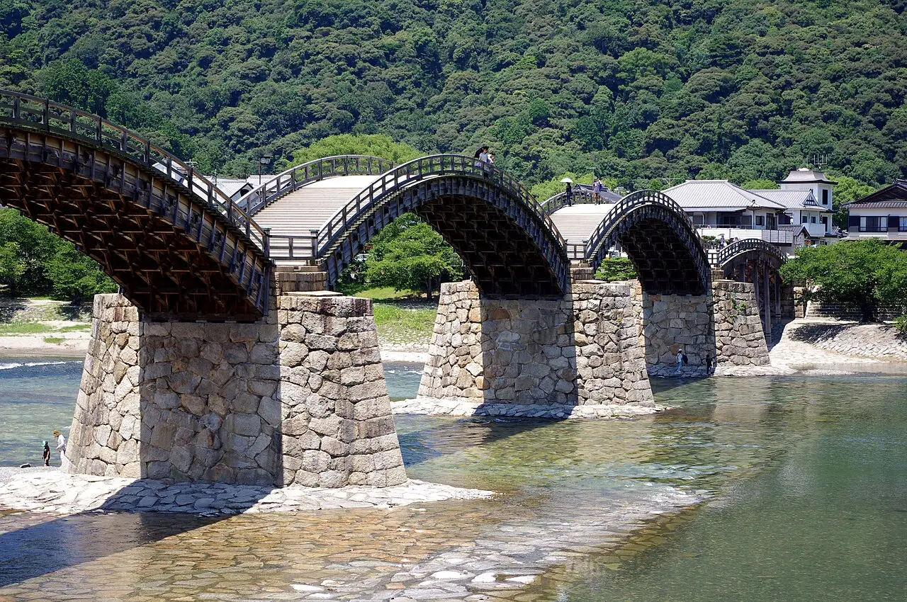 Ponte Kintai em Iwakuni, uma ponte de madeira em cinco arcos sobre pilares de pedra atravessando um rio raso, com uma encosta de mata ao fundo.