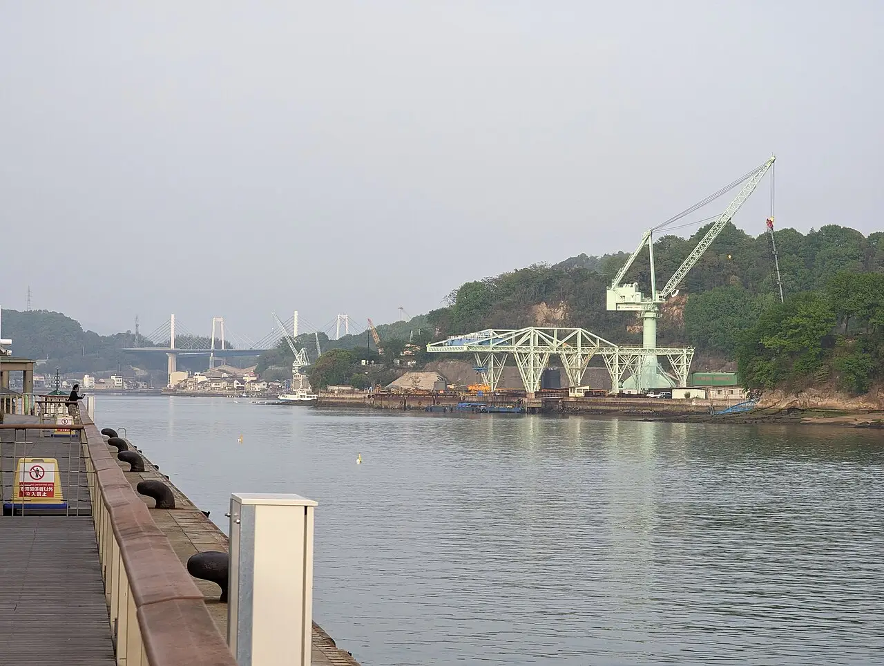 Estreito de Onomichi visto de um cais, com guindastes verdes de um estaleiro na margem oposta e uma ponte estaiada branca ao fundo.