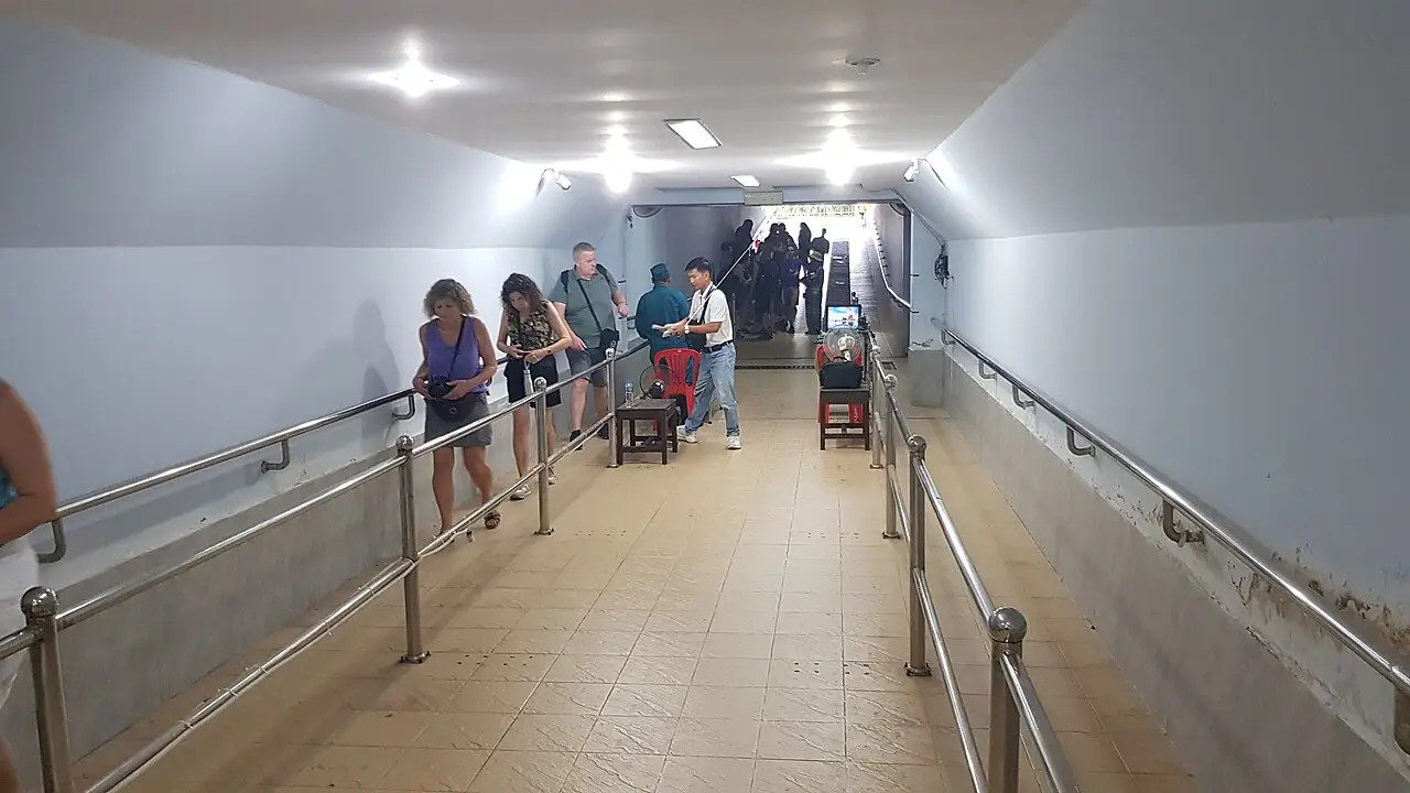 Corredor estreito com corrimãos e visitantes, descendo sob luzes no teto.