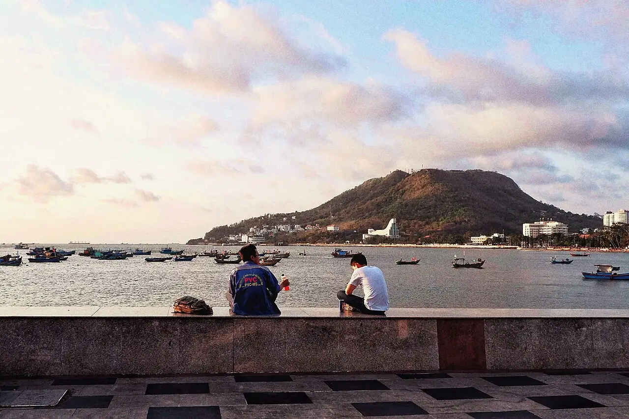 Baía de Vũng Tàu ao entardecer, com barcos de pesca ancorados, um morro ao fundo e dois homens sentados num muro à beira-mar.
