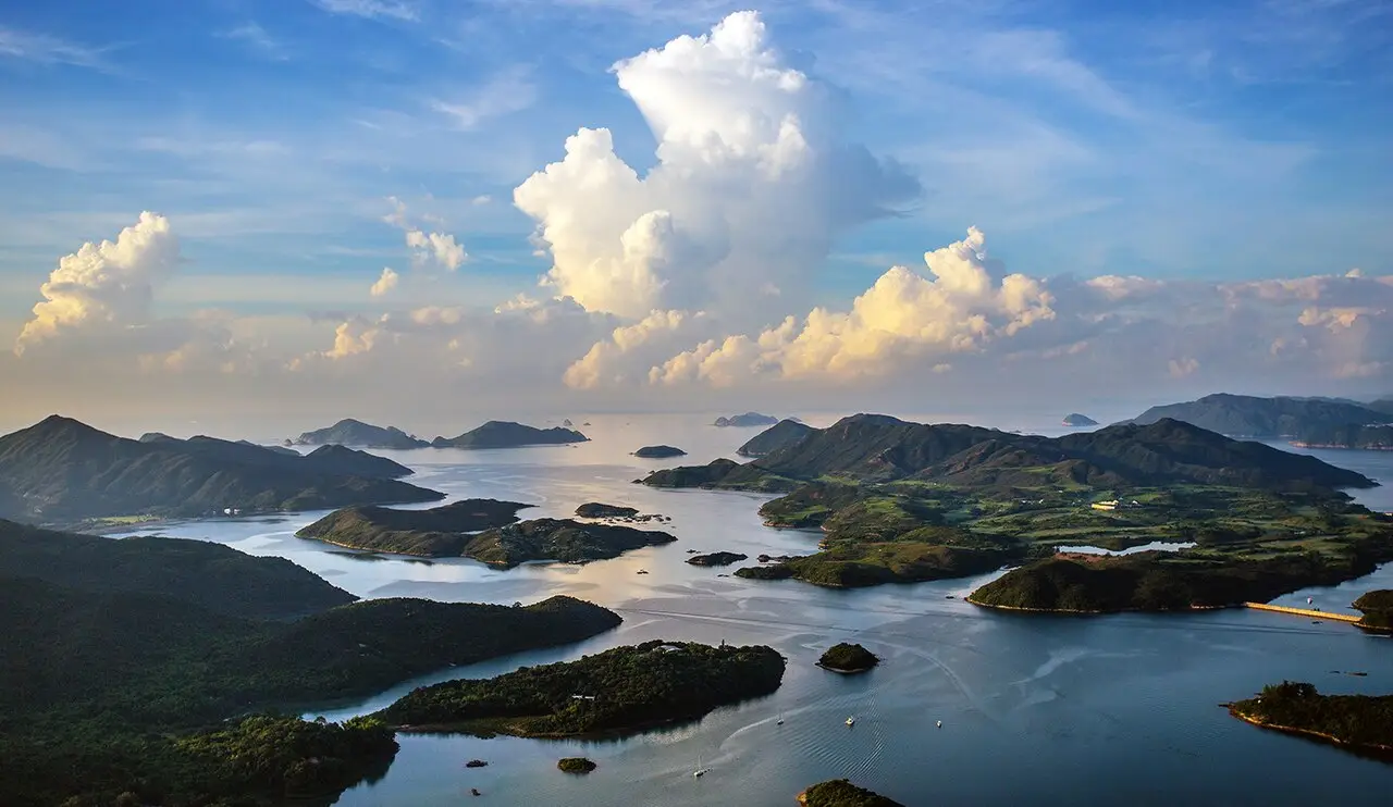 Vista do alto de ilhas verdes e enseadas de água calma em Sai Kung, com grandes nuvens iluminadas pelo sol baixo sobre o horizonte.