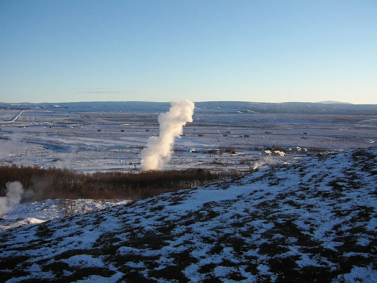 Coluna de vapor de um gêiser subindo numa planície coberta de neve, com casas pequenas ao lado e colinas geladas ao fundo.