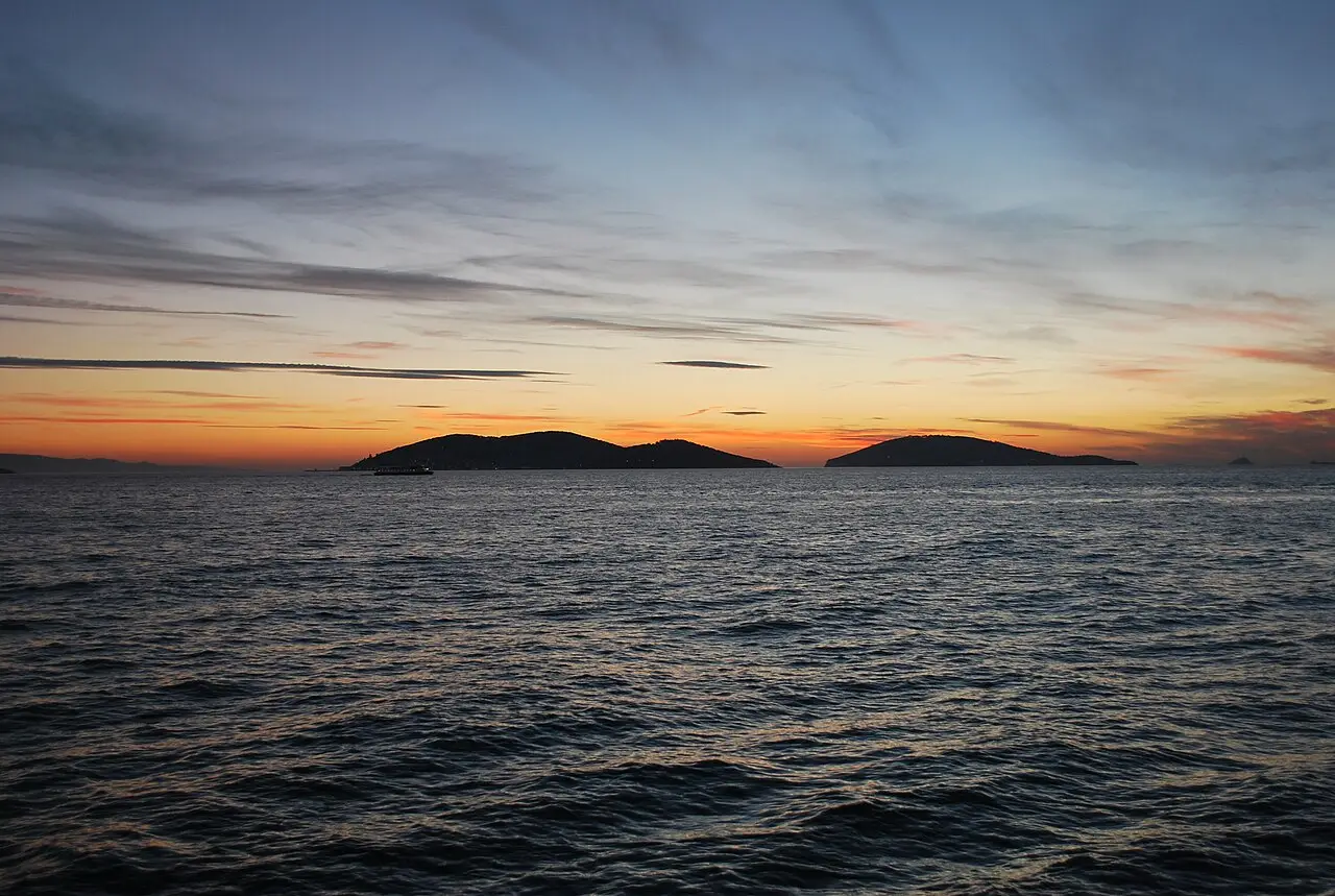 Silhueta de duas ilhas escuras no horizonte do mar de Mármara ao pôr do sol, com o céu alaranjado e azul e um pequeno barco à esquerda.