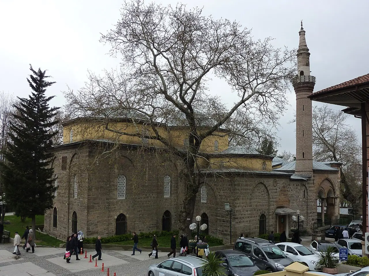 Mesquita de Orhan Gazi em Bursa, de pedra e tijolo com cúpulas amarelas e um minarete alto, atrás de um plátano sem folhas e de gente na praça.