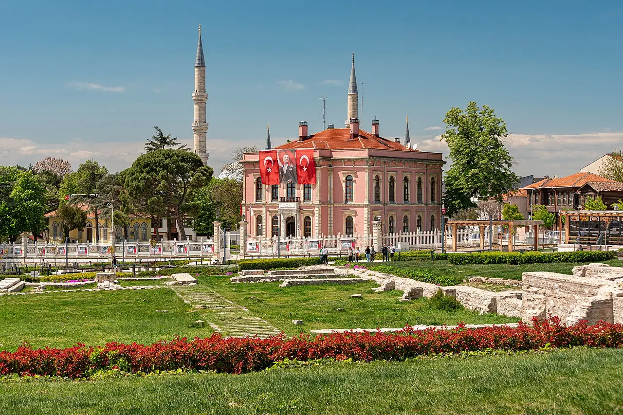 Edifício rosa de telhado de telhas com bandeiras turcas na fachada, dois minaretes ao fundo e ruínas de pedra em um jardim de canteiros vermelhos.
