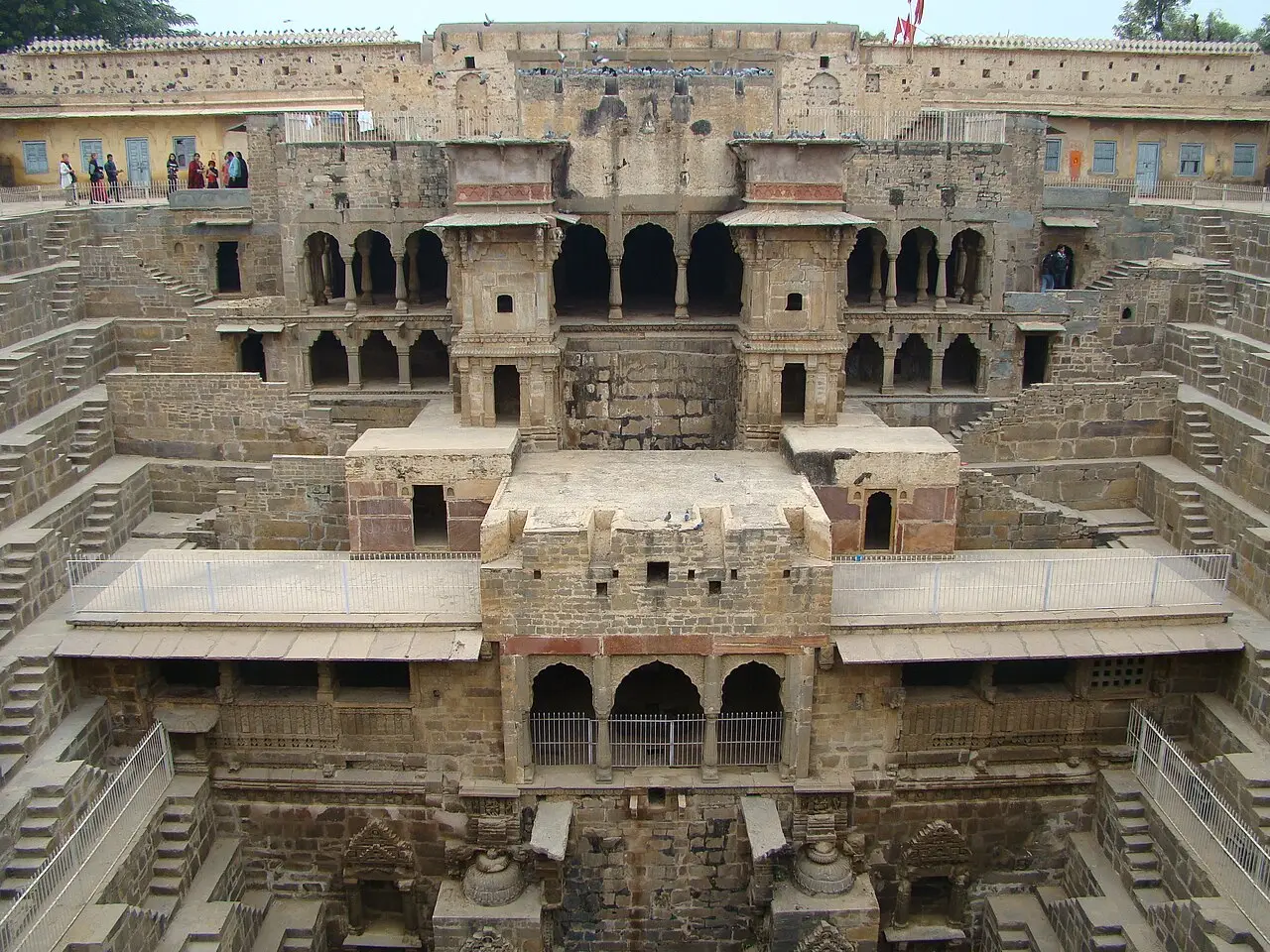 Poço escalonado Chand Baori visto de cima, com fileiras de degraus de pedra em todos os lados, galerias com arcos ao fundo e visitantes à esquerda.