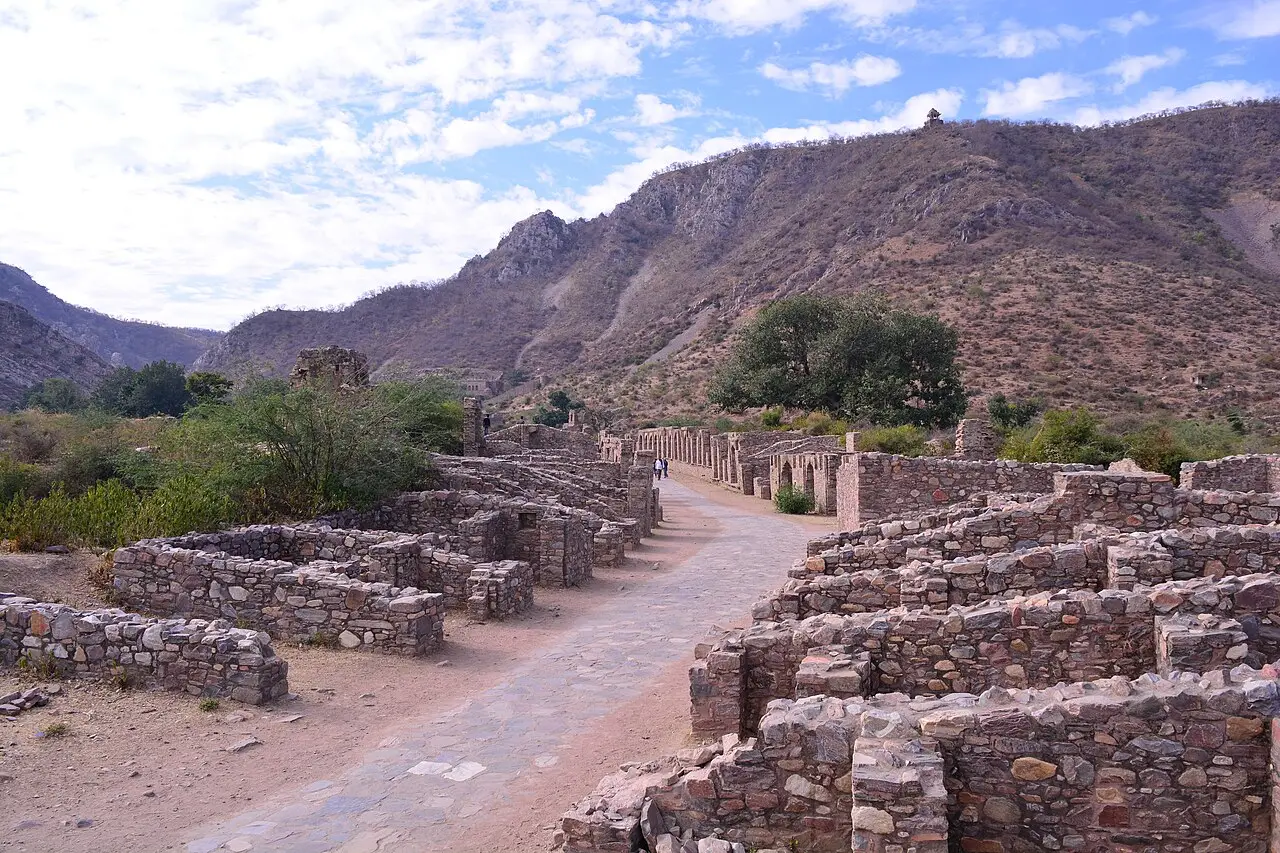 Ruínas de pedra de casas baixas ao longo de uma rua larga de pedra em Bhangarh, com colinas secas e um pequeno templo no topo ao fundo.