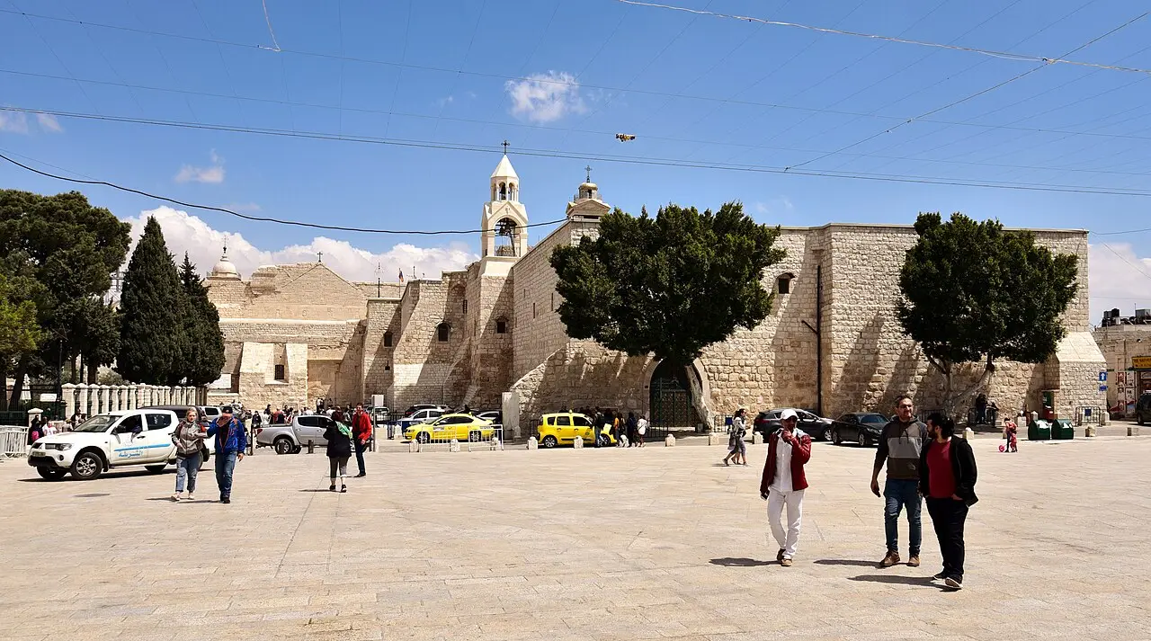 Fachada de pedra da Igreja da Natividade vista da Praça da Manjedoura, com árvores, veículos e visitantes sob céu azul.