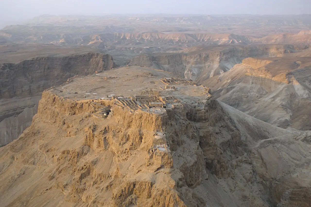 Vista aérea do platô de Massada, com ruínas sobre o topo plano de uma montanha de paredes íngremes e o deserto de vales secos ao redor.