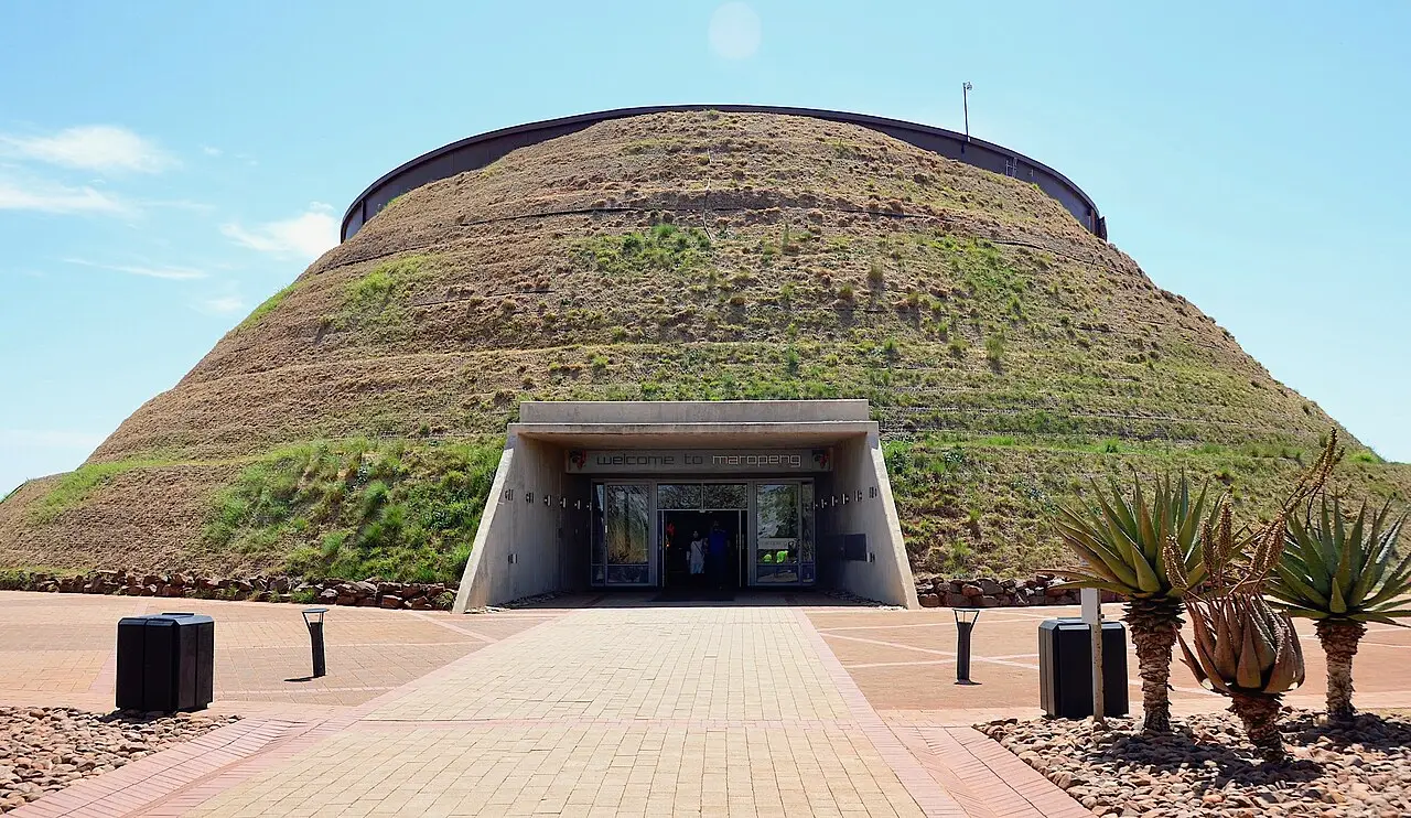 Entrada do centro de visitantes de Maropeng, num prédio em forma de montículo coberto de grama seca, com pavimento de tijolos e aloés em primeiro plano.