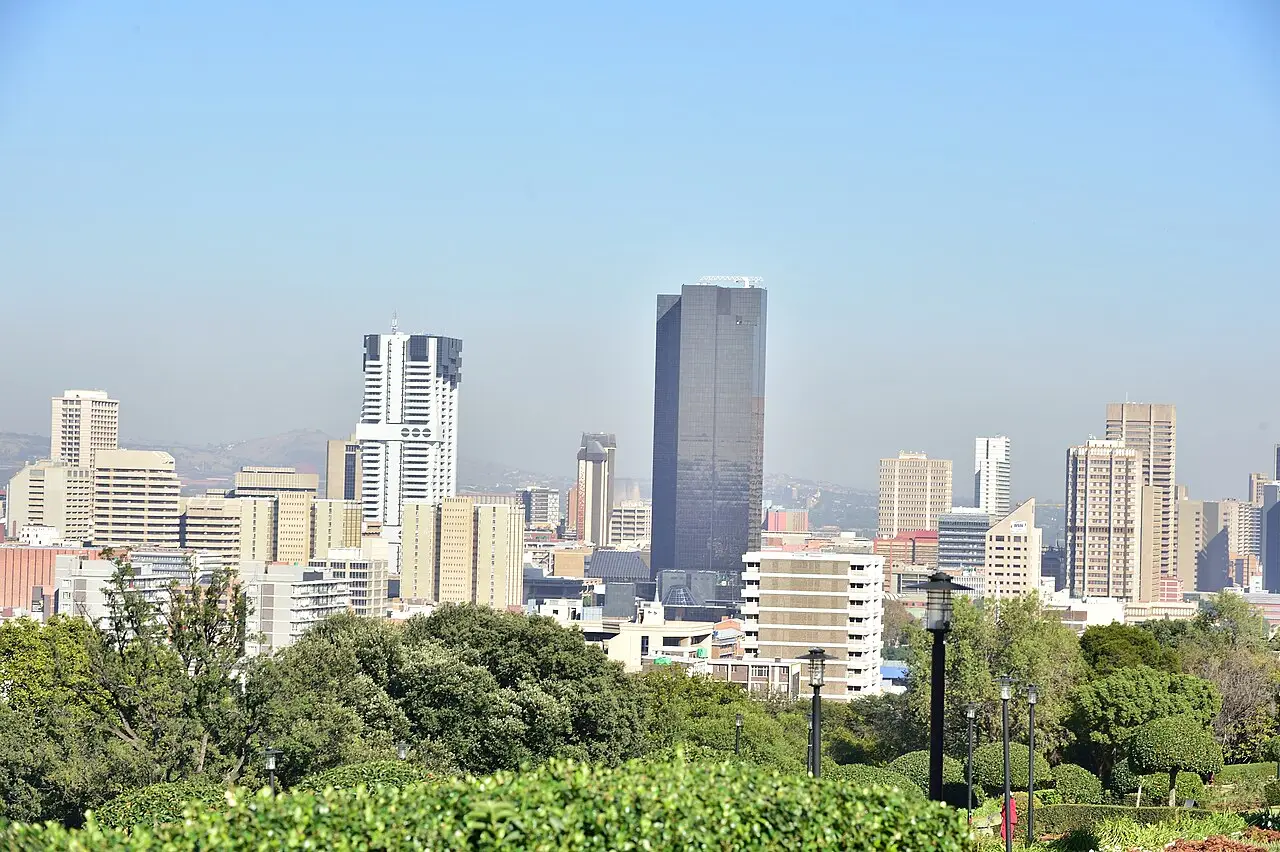 Vista de prédios altos do centro de Pretória entre a vegetação de um jardim em primeiro plano, sob céu azul com leve neblina.