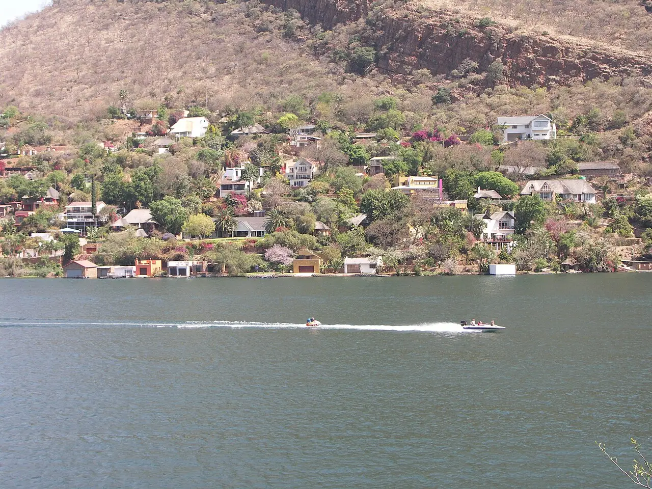 Casas espalhadas por uma encosta seca à beira da represa de Hartbeespoort, com uma lancha puxando um boia na água escura em primeiro plano.