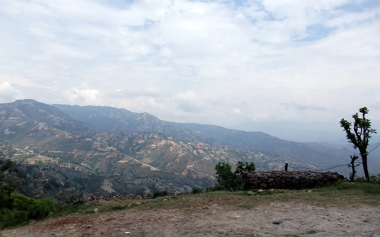 Colinas de encostas cultivadas em terraços perto de Dhulikhel, com um muro baixo de pedra e uma árvore à direita, sob céu de nuvens claras.