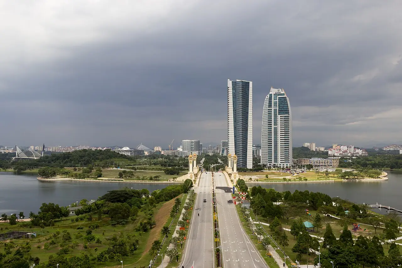 Ponte Seri Gemilang em Putrajaya, com uma avenida de duas pistas entre torres dourados, um lago dos dois lados e dois arranha-céus ao fundo sob céu carregado.