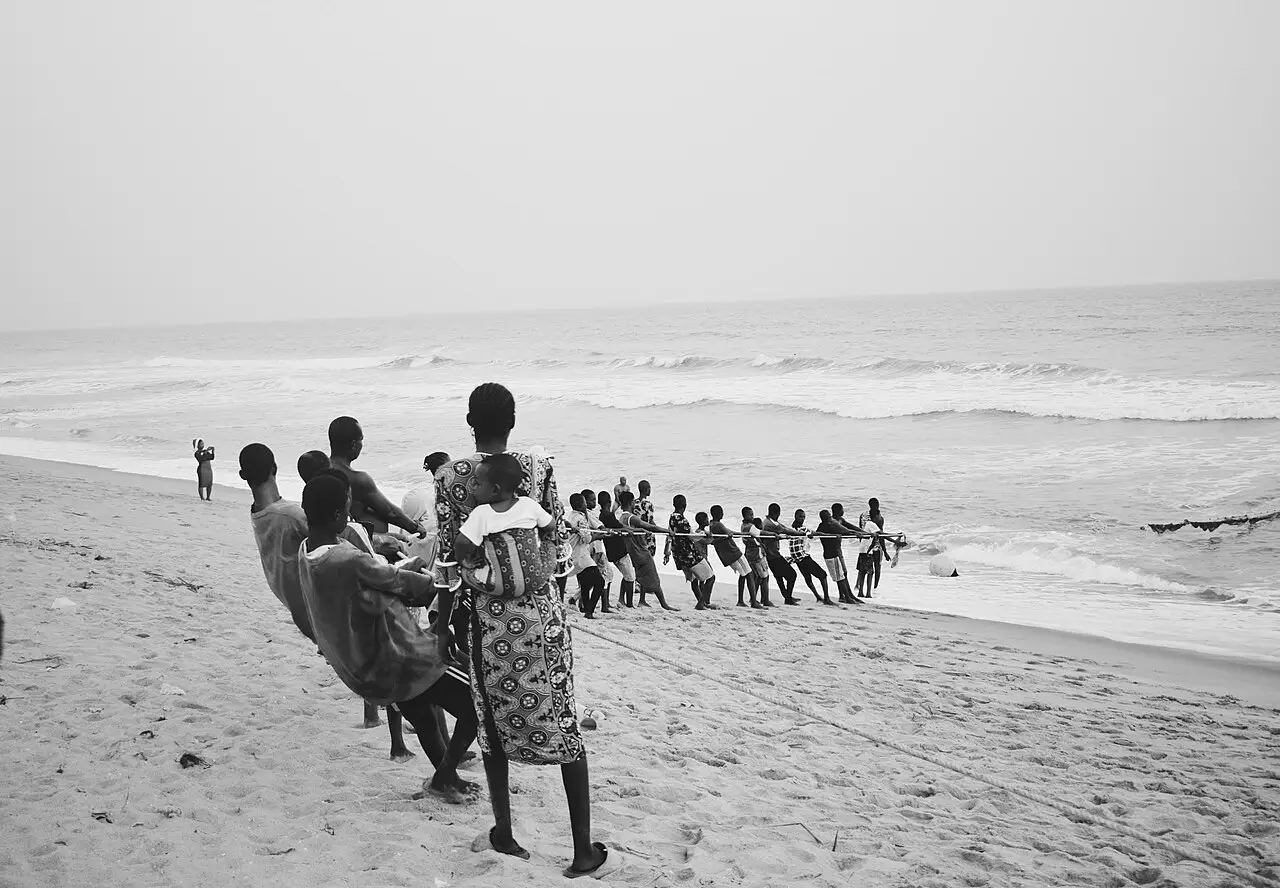 People haul a net on the sand, facing the sea, in a black and white photo.