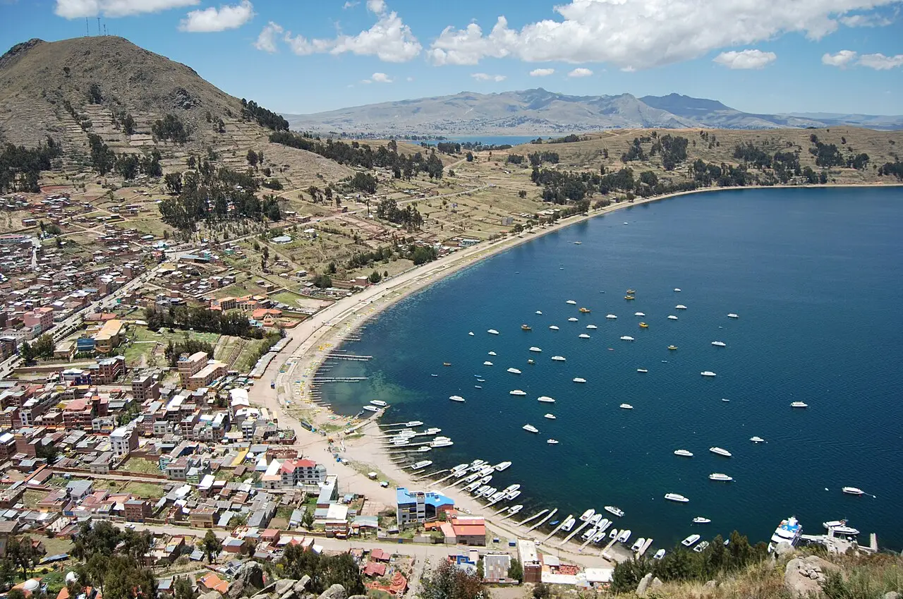 Vista do alto da baía de Copacabana, no lago Titicaca, com a cidade à esquerda, o cais com barcos e a água azul-escura cheia de embarcações brancas.