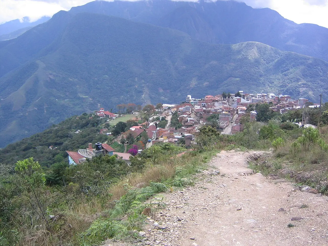 Vista de Coroico do alto de uma trilha de terra, com casas de telhado vermelho numa crista e montanhas íngremes cobertas de mata ao fundo.