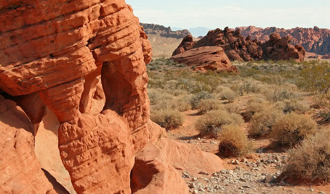 Rochas de arenito vermelho-alaranjado com camadas e buracos erodidos, e arbustos secos do deserto ao redor, com montanhas distantes ao fundo.
