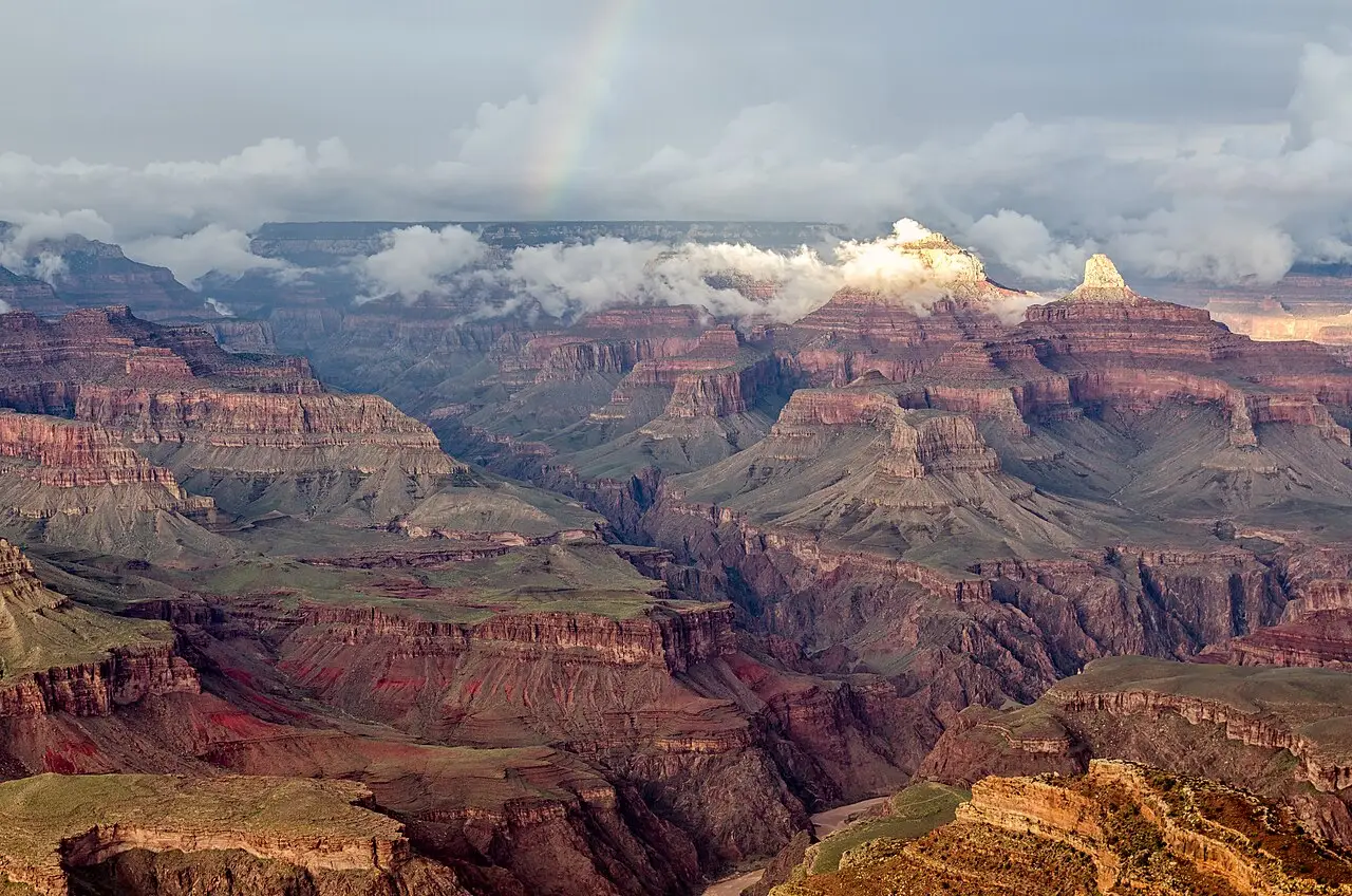Vista do Grand Canyon a partir de Hopi Point, com camadas de rocha vermelha, nuvens baixas entre os picos e um arco-íris no céu ao fundo.