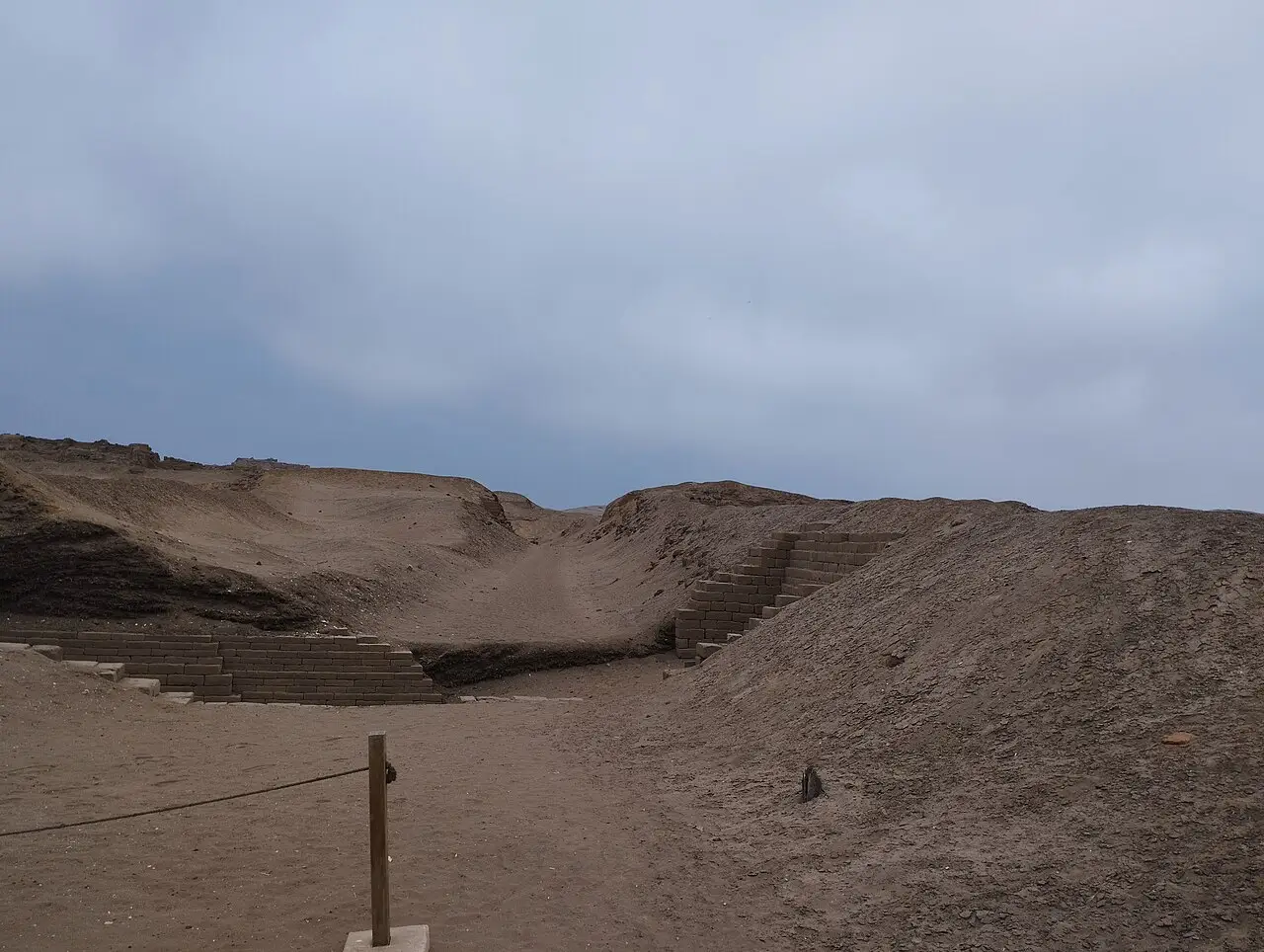 Muros de adobe em degraus num descampado de areia, sob céu cinza.