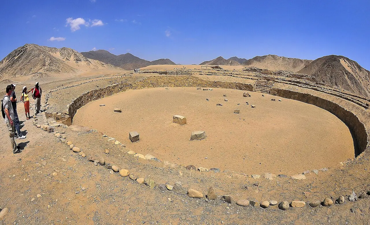 Praça circular afundada em Caral, com paredes de pedra baixas, blocos soltos no centro, um grupo de visitantes à esquerda e montanhas de deserto ao fundo.