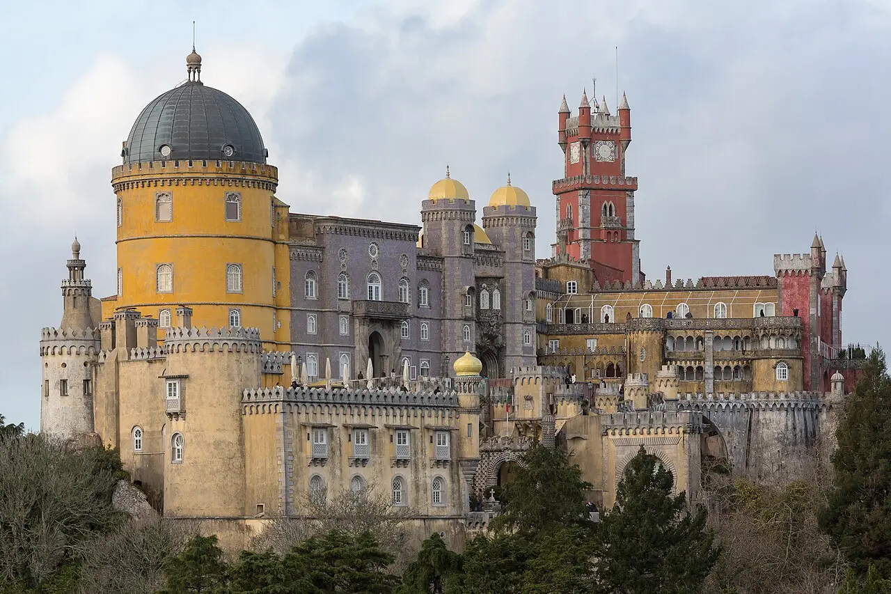 Palácio da Pena em Sintra, com a torre redonda amarela de cúpula escura, a torre do relógio vermelha, paredes ameadas e árvores em primeiro plano.