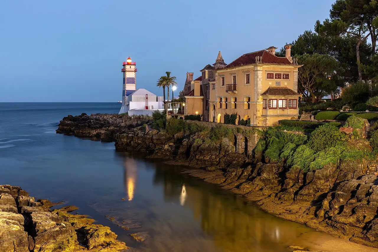 Farol de Santa Marta com listras azuis e brancas, aceso ao anoitecer, junto a uma casa de fachada amarela sobre rochas à beira do mar, em Cascais.