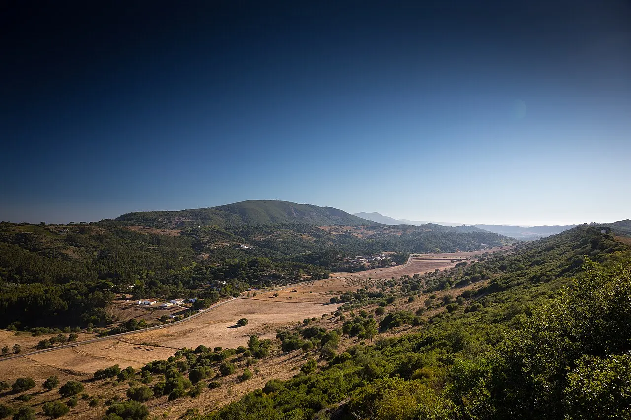 Colinas verdes e campos secos da Arrábida, com uma estrada, um povoado pequeno e o mar longe no horizonte.