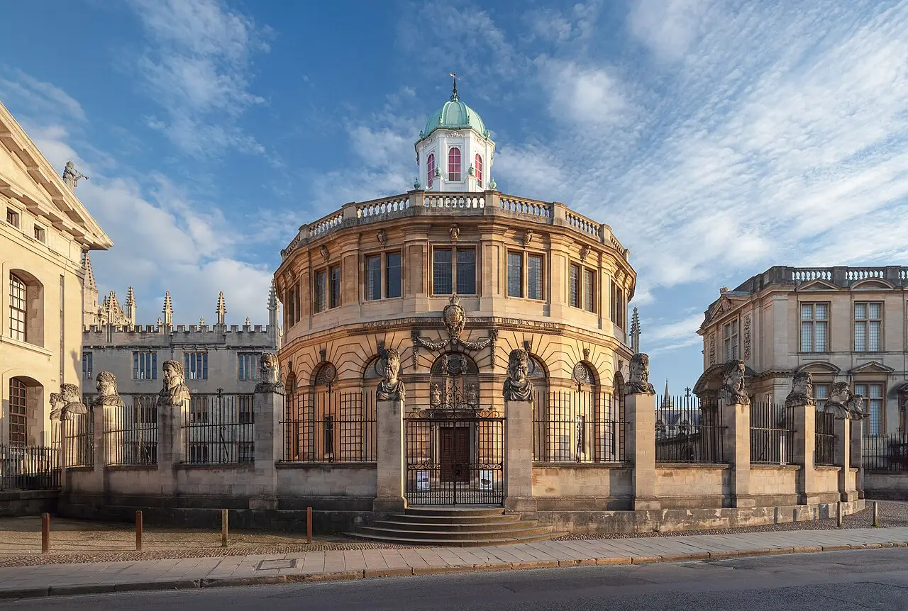 Fachada norte do Teatro Sheldonian em Oxford, de pedra clara e forma curva, com cúpula verde e uma fileira de bustos de pedra sobre o gradil de ferro.