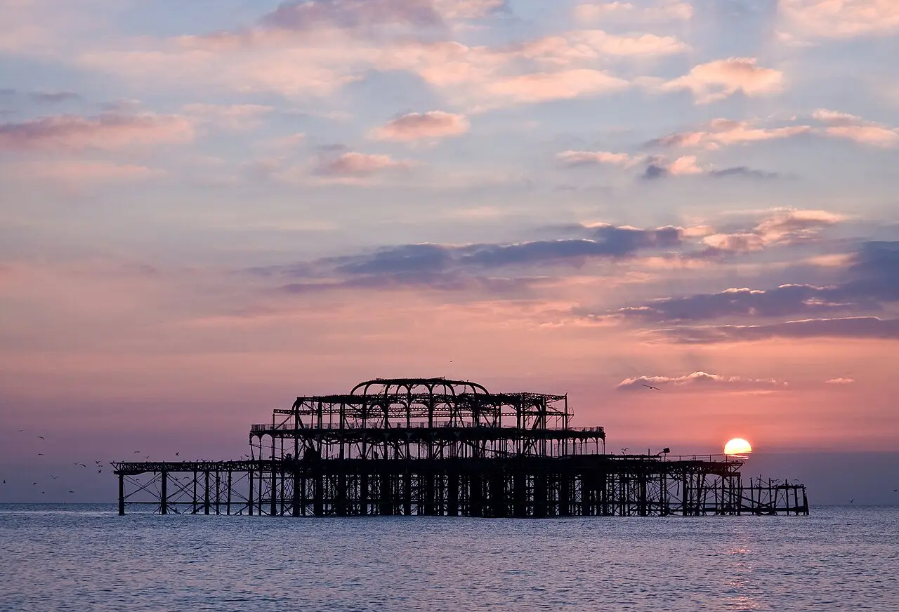 Estrutura de ferro em ruínas do West Pier de Brighton em silhueta no mar, com o sol se pondo à direita e um céu de nuvens cor de rosa.