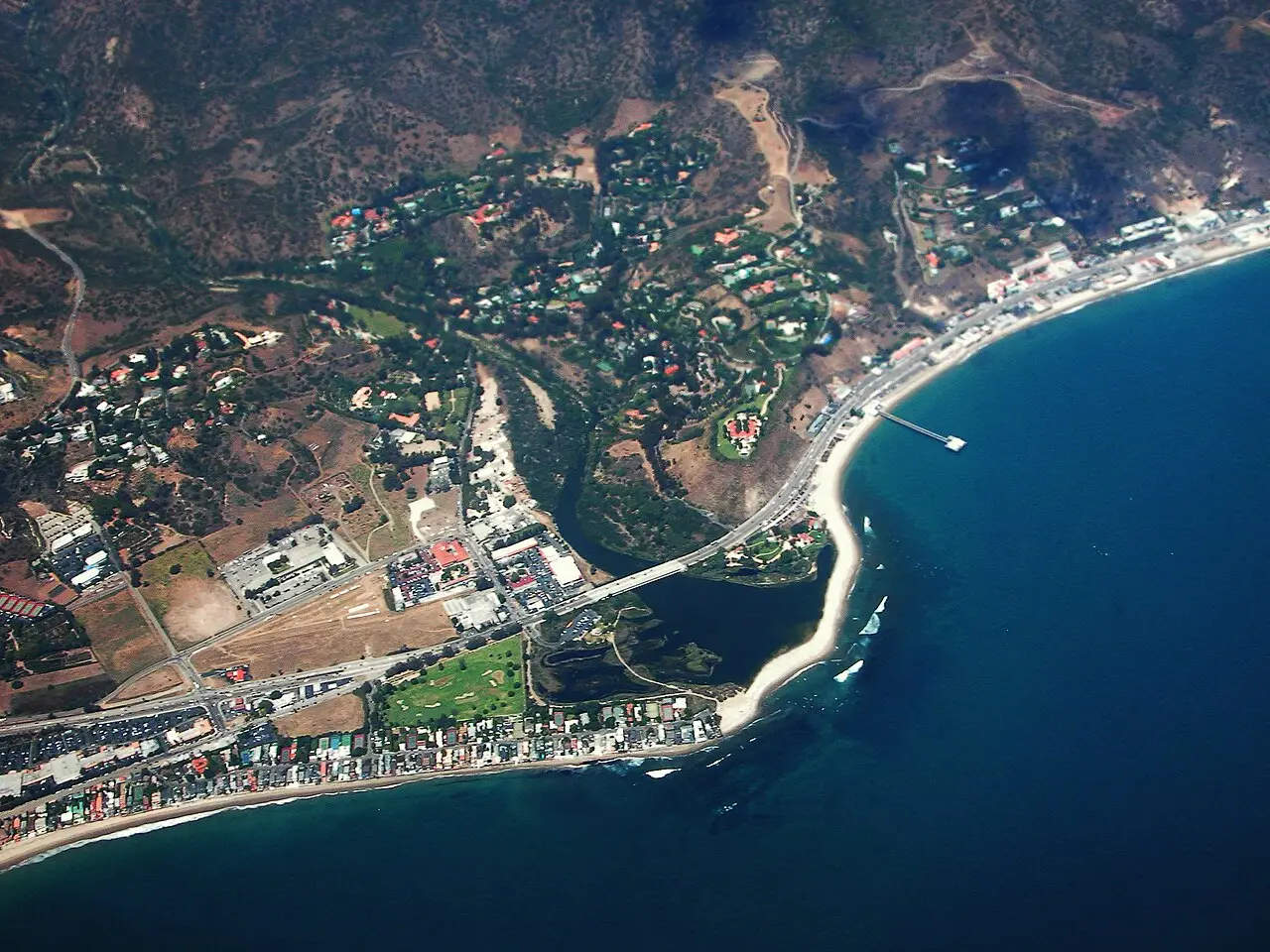 Vista aérea de Malibu, com a rodovia costeira, a laguna e o estuário atrás da faixa de areia, um píer à direita e o oceano azul-escuro em primeiro plano.
