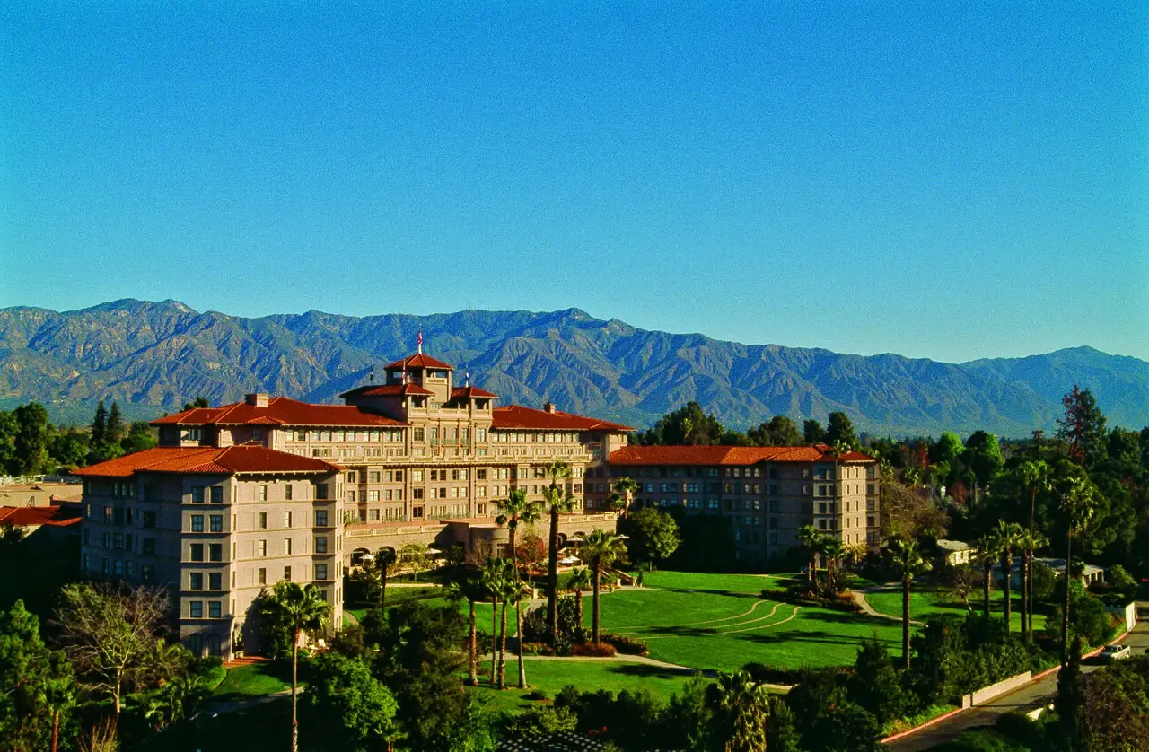 Hotel Langham Huntington em Pasadena, um edifício grande de telhas vermelhas cercado de gramado e palmeiras, com montanhas ao fundo sob céu azul limpo.