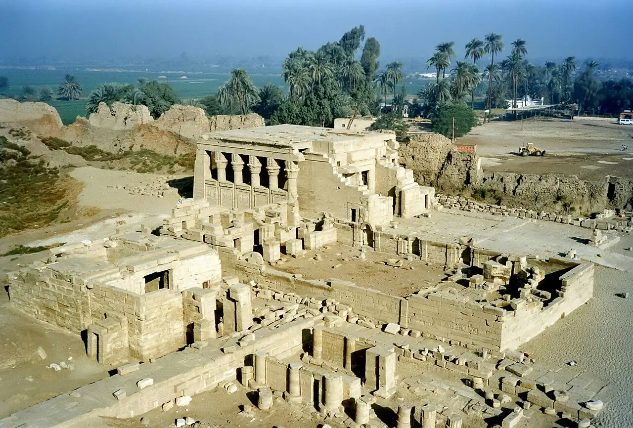 Vista de cima de parte do complexo do templo de Dendera, com um pórtico de colunas, muros de pedra e ruínas, e palmeiras e campos verdes ao fundo.