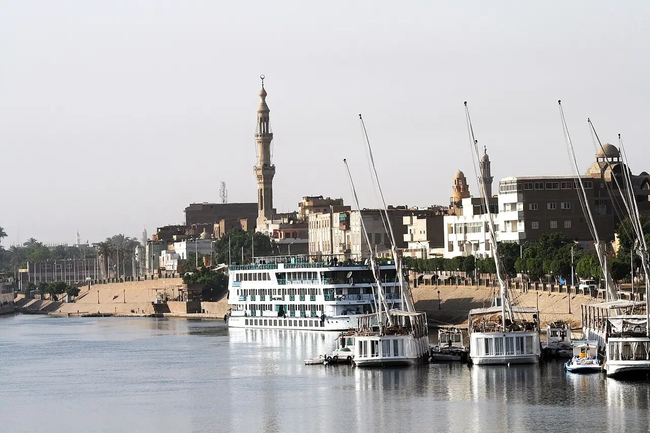 Margem do Nilo em Esna, com barcos de cruzeiro brancos e feluchas atracados, um minarete alto e prédios baixos da cidade ao fundo.