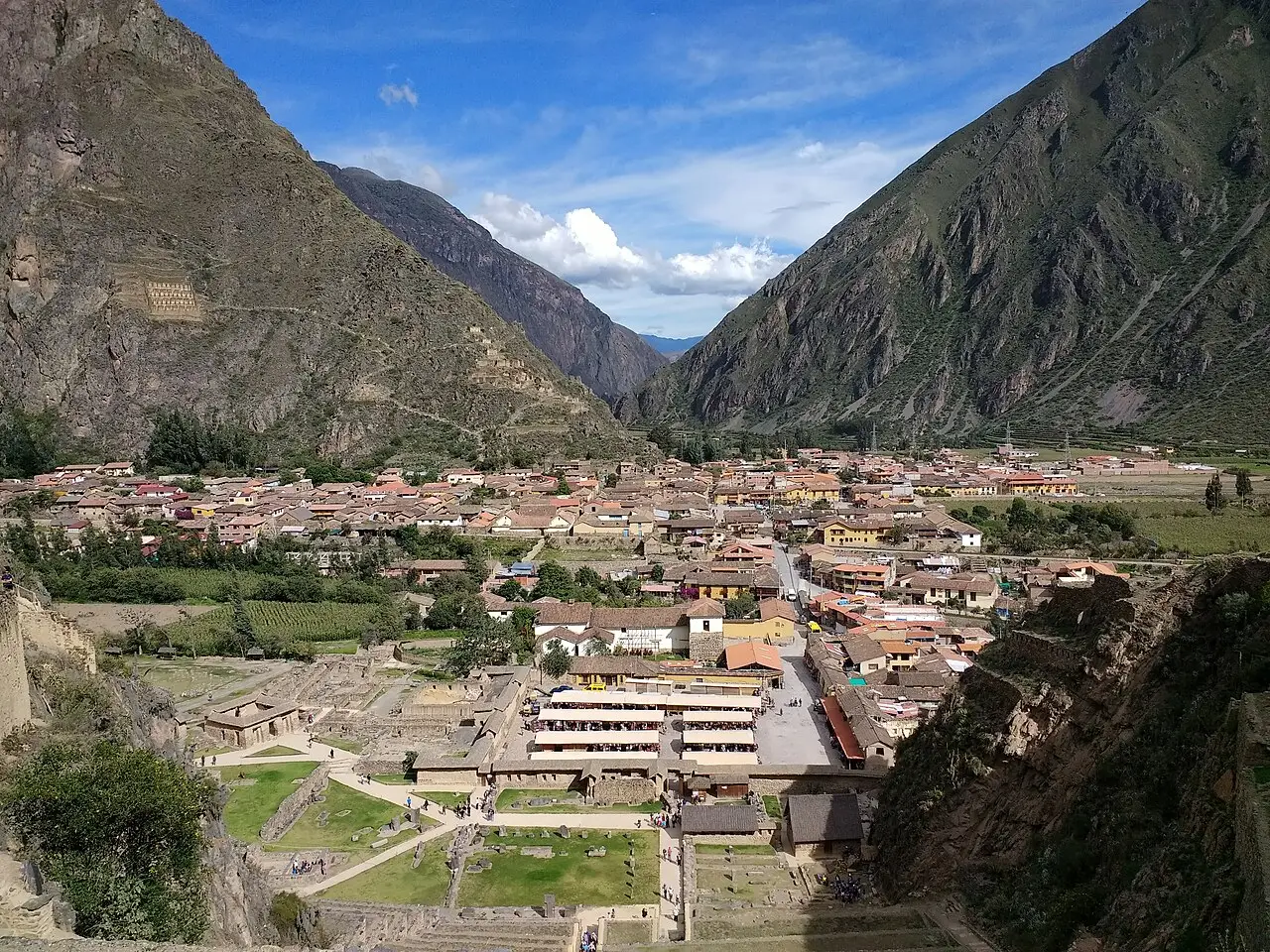 Vista alta de Ollantaytambo, com os telhados do povoado no vale, as ruínas com terraços de pedra em primeiro plano e montanhas íngremes dos dois lados.