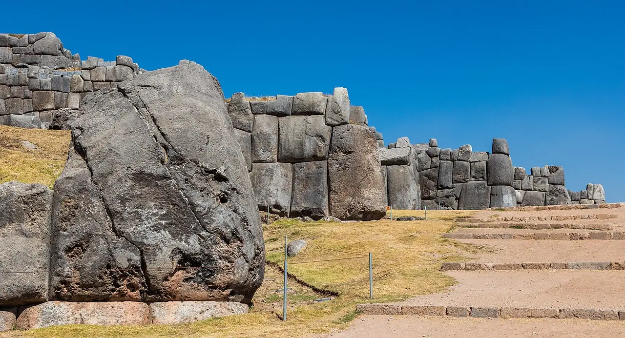 Muralha de blocos de pedra enormes e encaixados em Sacsayhuamán, com uma rocha grande em primeiro plano, degraus de terra à direita e céu azul limpo.