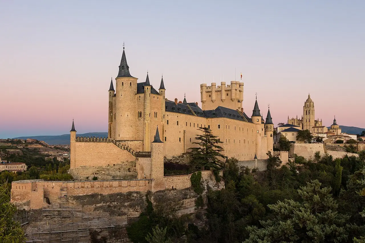 O Alcázar de Segovia ao entardecer, com torres de telhado pontudo sobre uma rocha, muralhas de pedra clara e a catedral ao fundo à direita.