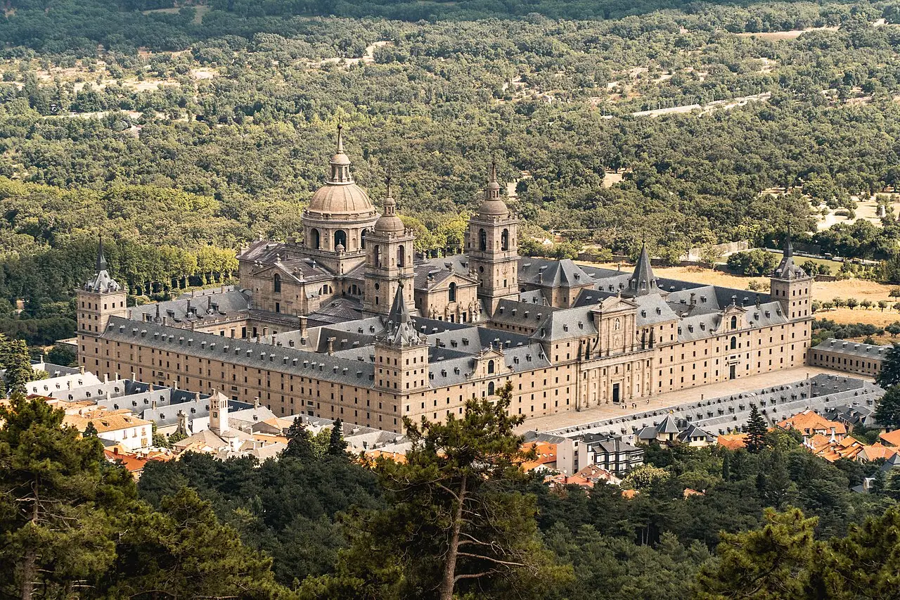 Vista aérea do mosteiro de El Escorial, um bloco grande de pedra clara com cúpula e torres, cercado de floresta e com casas da vila junto ao pé.