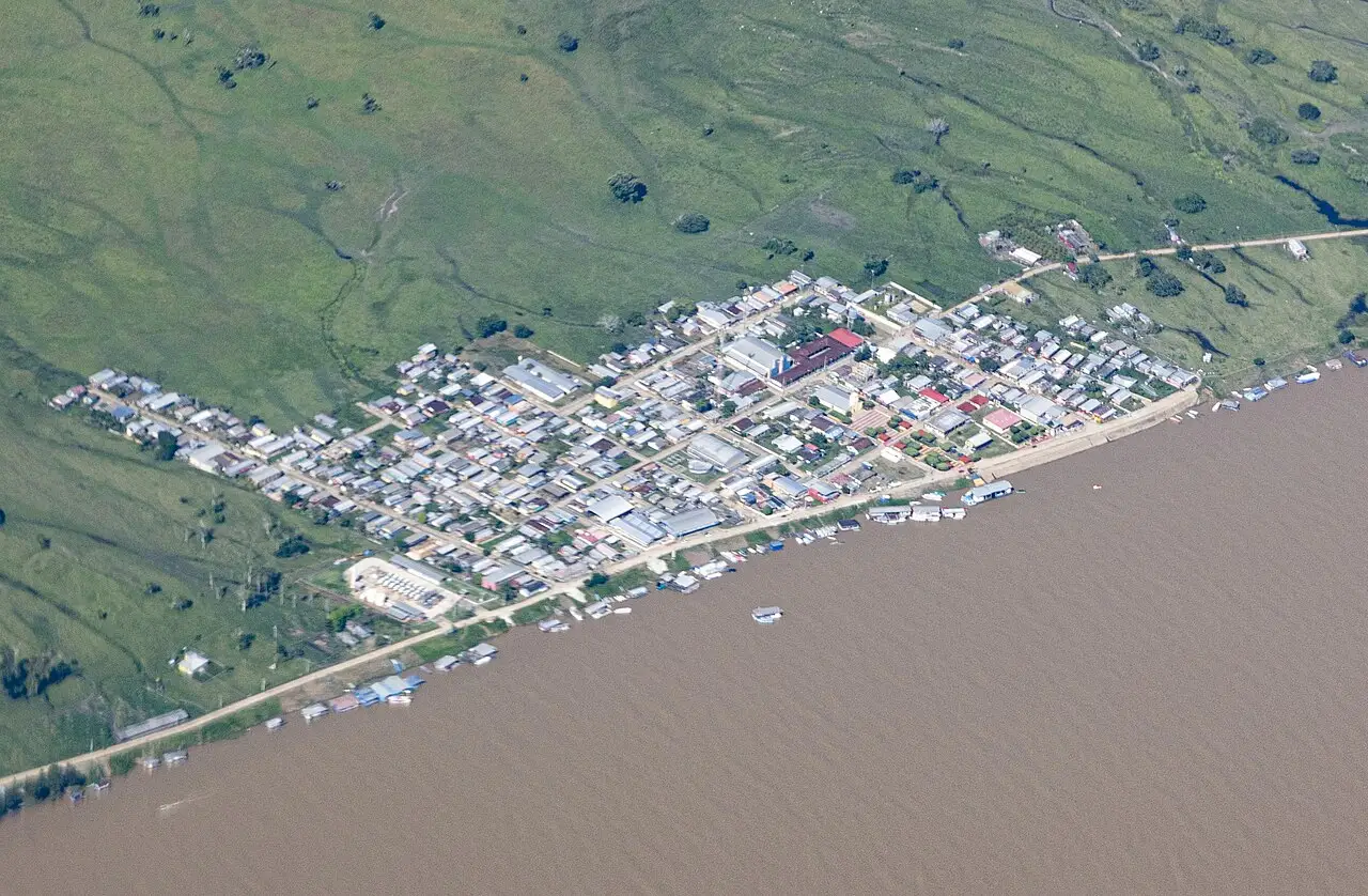 Pueblo a orillas de un río lodoso, con casas, barcos y campo verde, visto desde arriba.