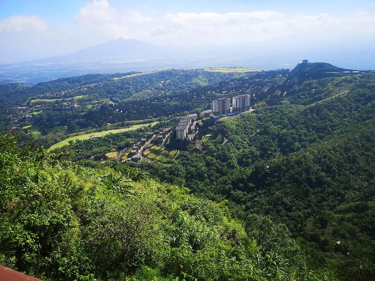 Vista de cima das terras altas de Tagaytay, com encostas cobertas de mata, um campo de golfe, prédios residenciais e uma montanha distante sob nuvens.