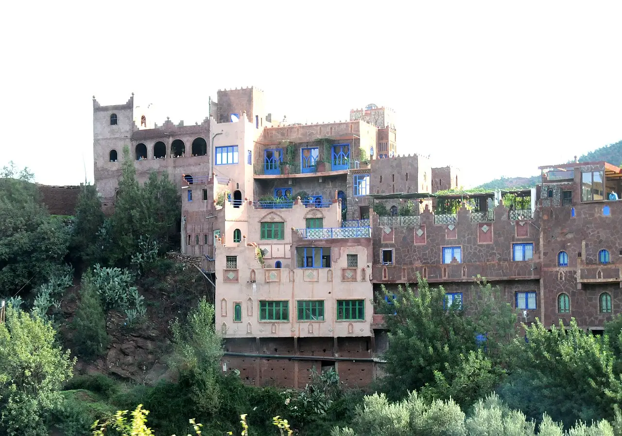 Edifício de vários andares em tons de rosa e terracota com janelas azuis e verdes, torres com ameias e árvores em volta, numa encosta do vale do Ourika.