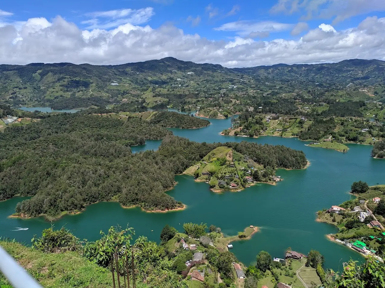 Vista do alto sobre o reservatório de Guatapé, com braços de água verde-azulada entre penínsulas cobertas de mata, casas na margem e colinas ao fundo.
