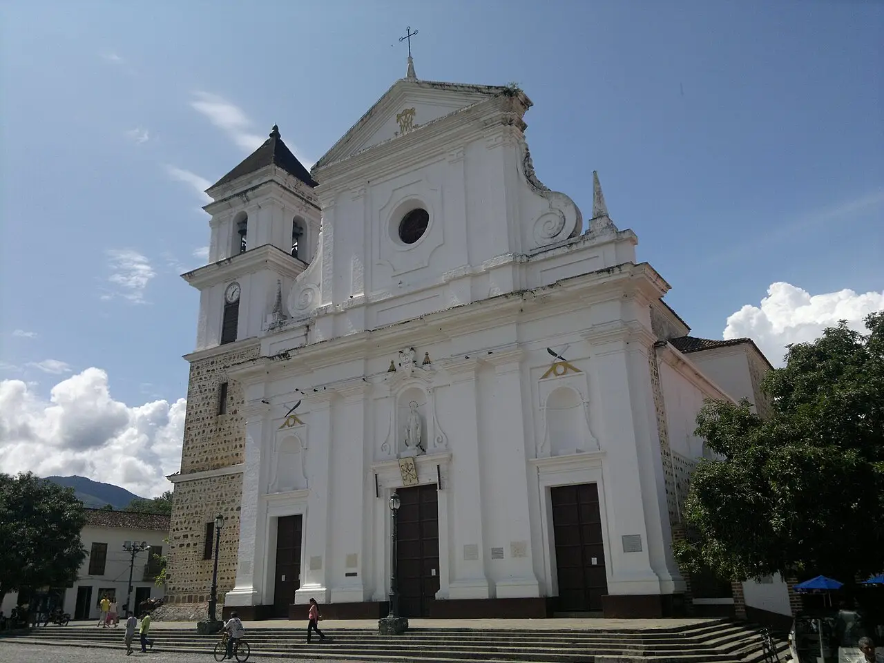 Fachada branca da catedral de Santa Fe de Antioquia, com torre de relógio, três portas de madeira, escadaria e a praça em frente.